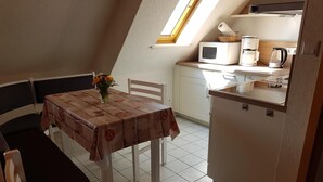 Pieni, käytännöllinen keittiö, jossa on mikroaaltouuni, kahvinkeitin ja vedenkeitin. Keittiössä on ruokapöytä, jonka päällä on kukkakoriste, sekä yksi tuoli. Huoneessa on kalteva katto ja kattovalokuvio.