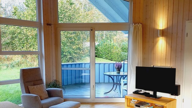 Ein gemütliches Wohnzimmer mit Fernseher, einem bequemen Sessel, einem kleinen Tisch und Blick auf den Garten durch große Fenster.