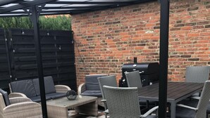 Eine Terrass mit Backsteinwand, einer Pergola, Korbmöbeln und einem Grill.