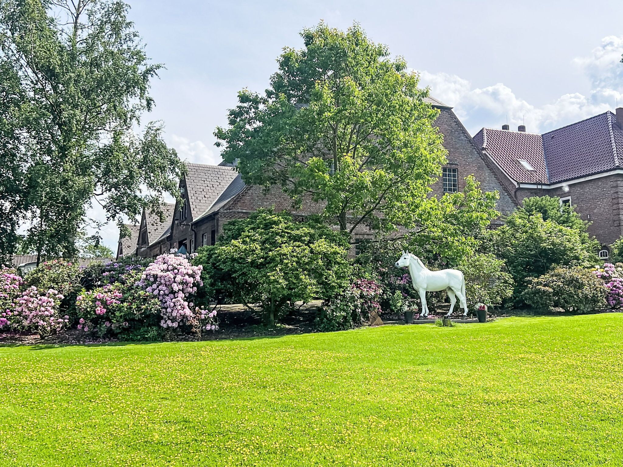 A grassy lawn with a white horse statue, surrounded by trees and shrubs, with a building in the background.