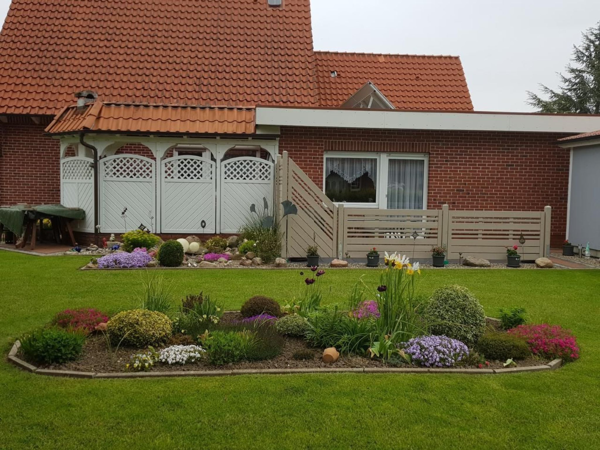 A two-story brick house with a red-tiled roof, a front porch with a bench, and a well-maintained garden with various flowers and shrubs.