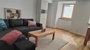 A modern living room with a grey sofa, black ottoman, wooden coffee table, and a large window with blinds. - Apartment Günter by Interhome (Sagogn)