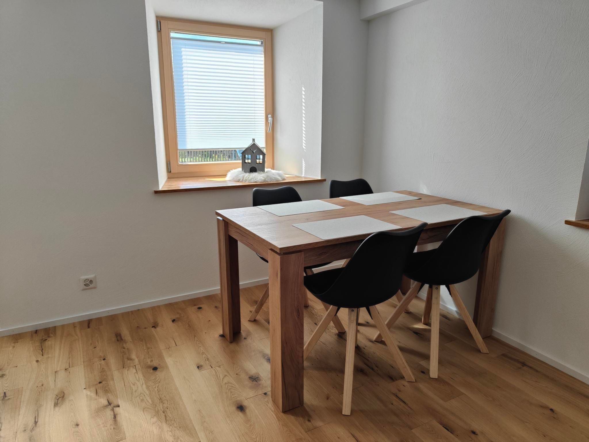 A dining area with a wooden table and black chairs, a window with blinds, and a small house decoration on the windowsill.