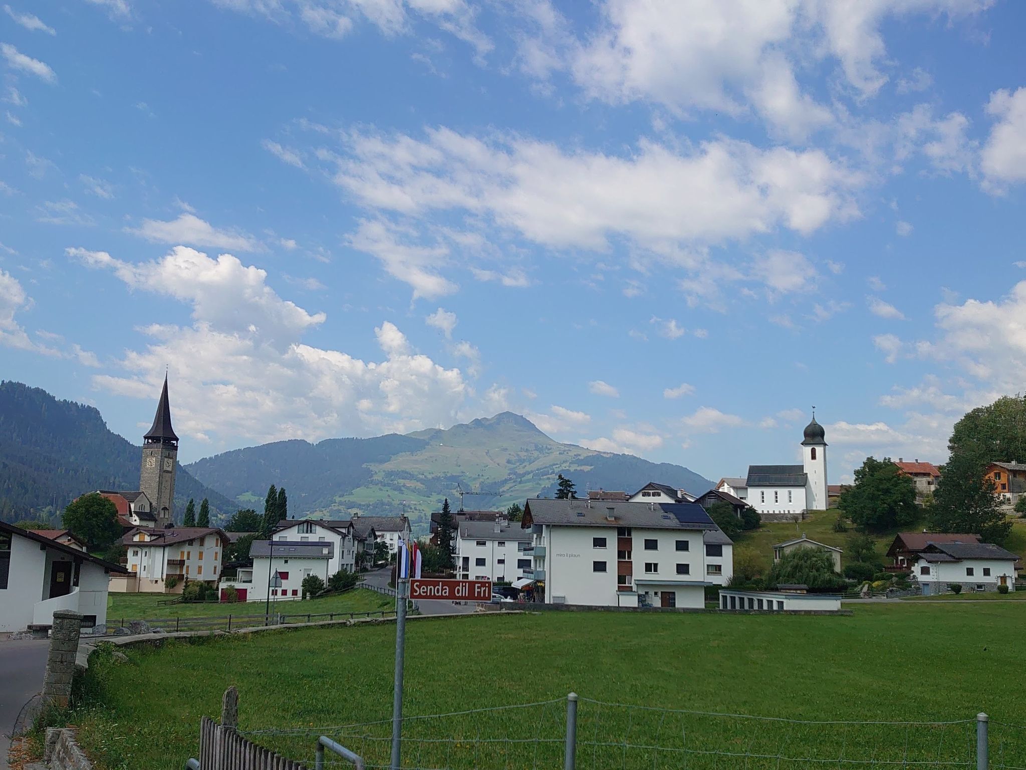 A town with a church, a sign reading "Senda di Fri", and houses in a valley with mountains in the background.