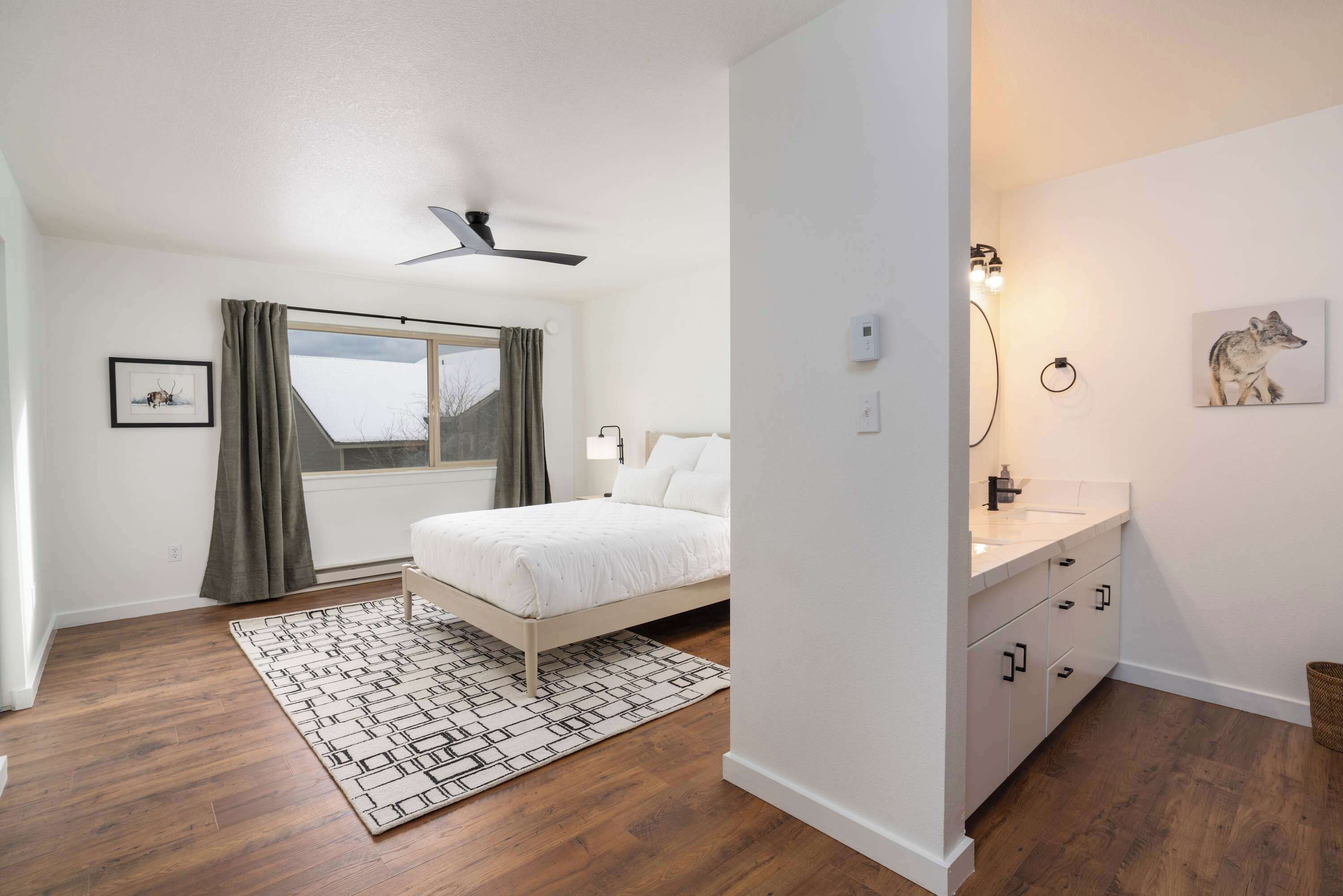 Un dormitorio con cama, ventilador de techo, ventana con cortinas, mesita de noche y lámpara de pared.