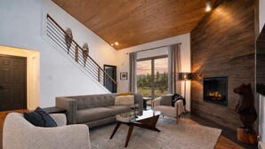 A modern living room with a staircase, a fireplace, and a large window with a view. - The Drift at Firelight | Big Sky Adventure+Hot Tub (Big Sky)