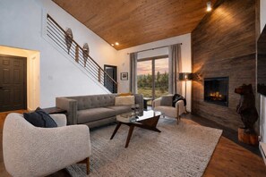 A modern living room with a staircase, a fireplace, and a large window with a view. - The Drift at Firelight | Big Sky Adventure+Hot Tub (Big Sky)
