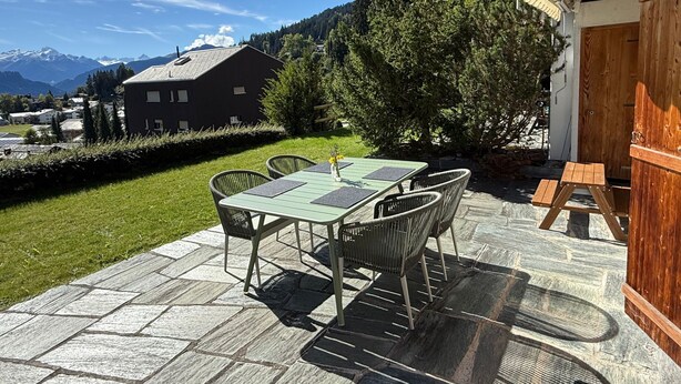 An outdoor patio with a table and chairs, a wooden bench, and a view of a mountain and forest. - Familienfreundliche und gemütliche 3.5 Wohnung mit Garten by Interhome (Laax)