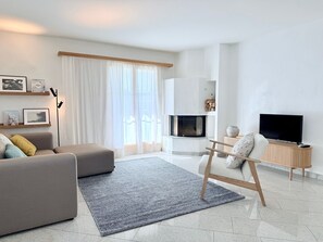 A modern living room with a sofa, a TV, a fireplace, and a large window with sheer curtains. - Familienfreundliche und gemütliche 3.5 Wohnung mit Garten by Interhome (Laax)