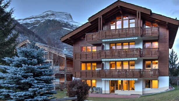 A multi-story wooden building with balconies, surrounded by snow-covered mountains and evergreen trees. - Top Ferienwohnung Breithorn (SkiIn/SkiOut) by Interhome (Zermatt)