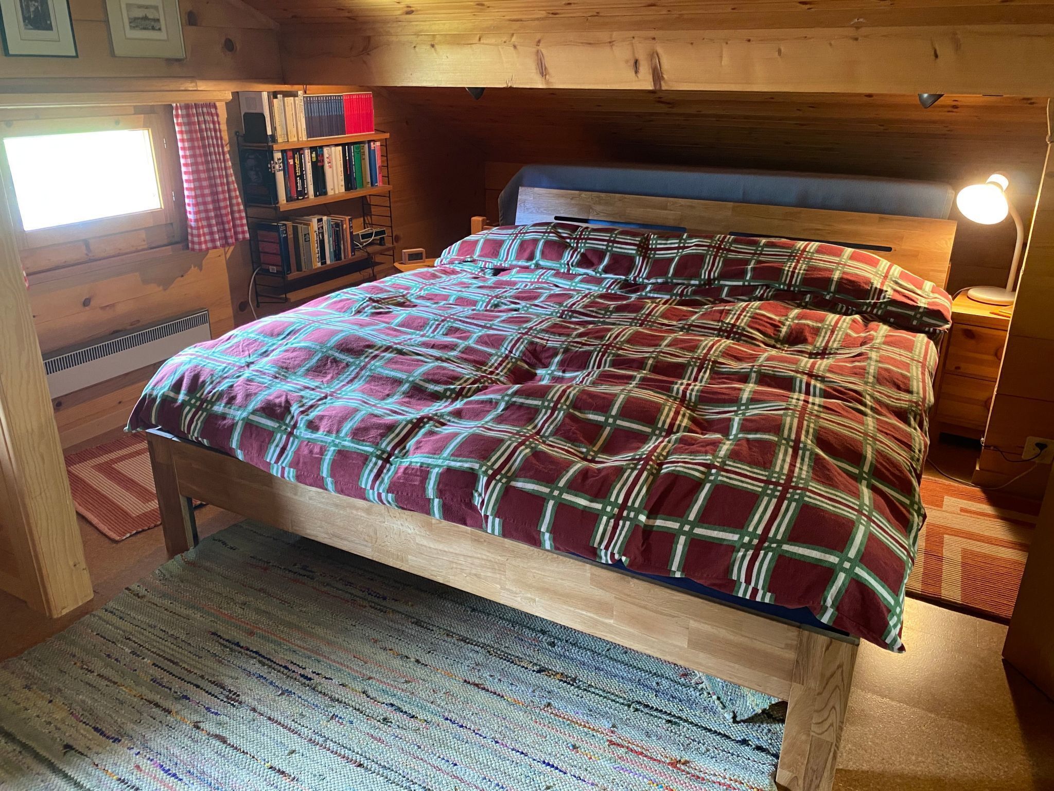 Une chambre dans une cabane en bois, avec une couverture à carreaux sur le lit, une bibliothèque et une fenêtre avec des rideaux.