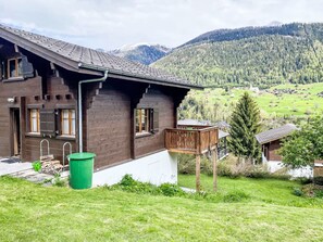 Una cabaña de madera con balcón, un contenedor verde para basura y vistas de un paisaje montañoso.