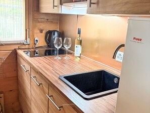 Cocina compacta con muebles de madera, un lavamanos negro, una tetera y una botella de vino.