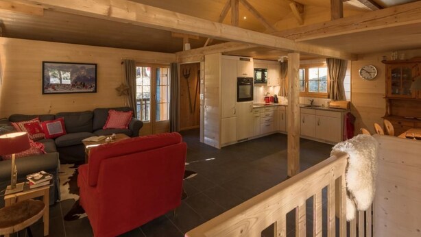 Un amplio salón con un sofá rojo, un sofá negro y una mesa de comedor de madera. Hay una zona de cocina con armarios blancos y un reloj en la pared.
