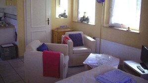 A small, well-lit room with a sofa, a chair, a small table, and a television. There are two windows with curtains, a door, and a mirror on the wall. - Strandfloh in Hörnum by Interhome (Hörnum)