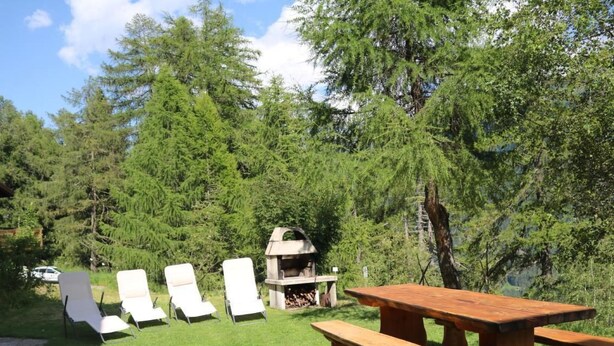An outdoor seating area with a wooden picnic table and white chairs, surrounded by lush greenery and trees. - Chalet Männertreu OG by Interhome (Bellwald)
