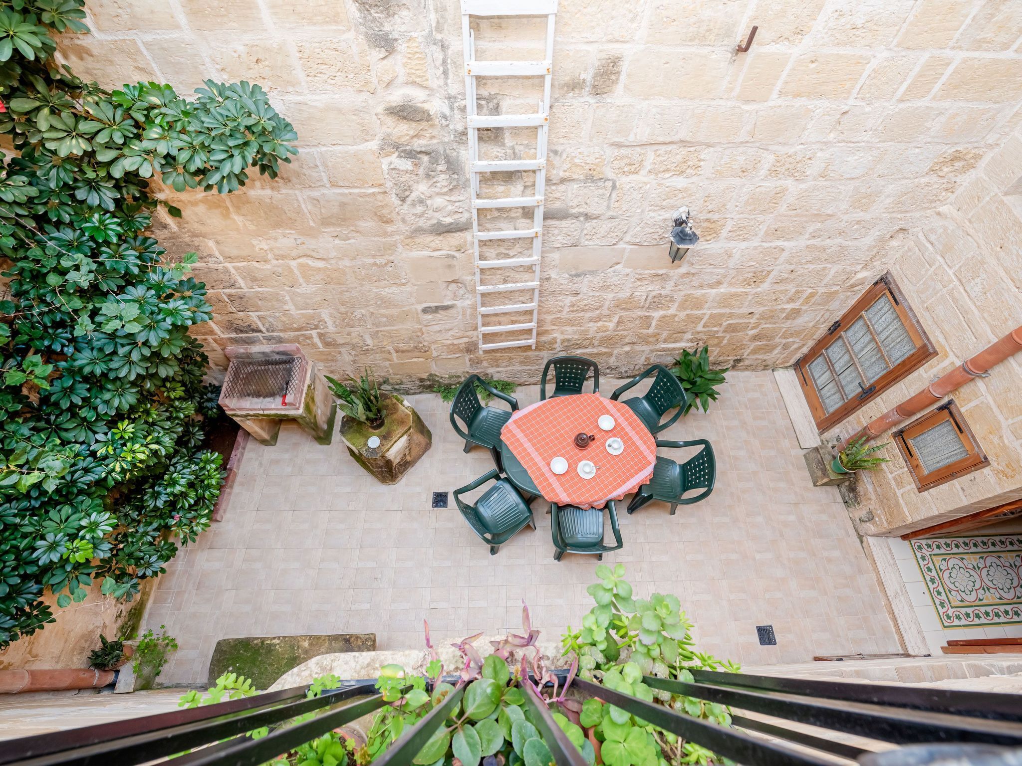 A stone courtyard with a round table and chairs, surrounded by plants and a ladder.