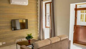 Ett vardagsrum med en soffa, en tv som är monterad på en vägg av trä, en krukväxt och ett fönster med gardiner.