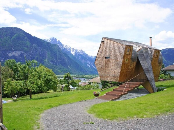 Una casa con un originale tetto spiovente, immersa in una zona montuosa e raggiungibile tramite un vialetto sterrato.
