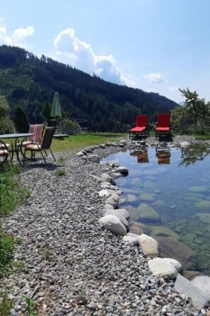 A clear outdoor area with a pebble path leading to a calm water body, surrounded by a forested hill. There are red chairs and a table with umbrellas, suggesting a seating area for relaxation.