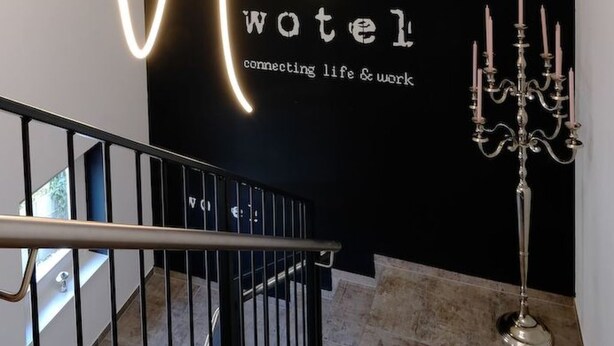 Ein modernes Hotel-Interieur mit einer Treppe, einer schwarzen Wand mit der Aufschrift 'hotel' und 'verbindet Leben & Arbeit' sowie einem Kandelaber.