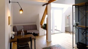 Ein geräumiges Loft mit freiliegenden Holzbalken, einem Essbereich mit Tisch und Stühlen, einem farbenfrohen Teppich und einem großen Fenster, das für natürliches Licht sorgt.