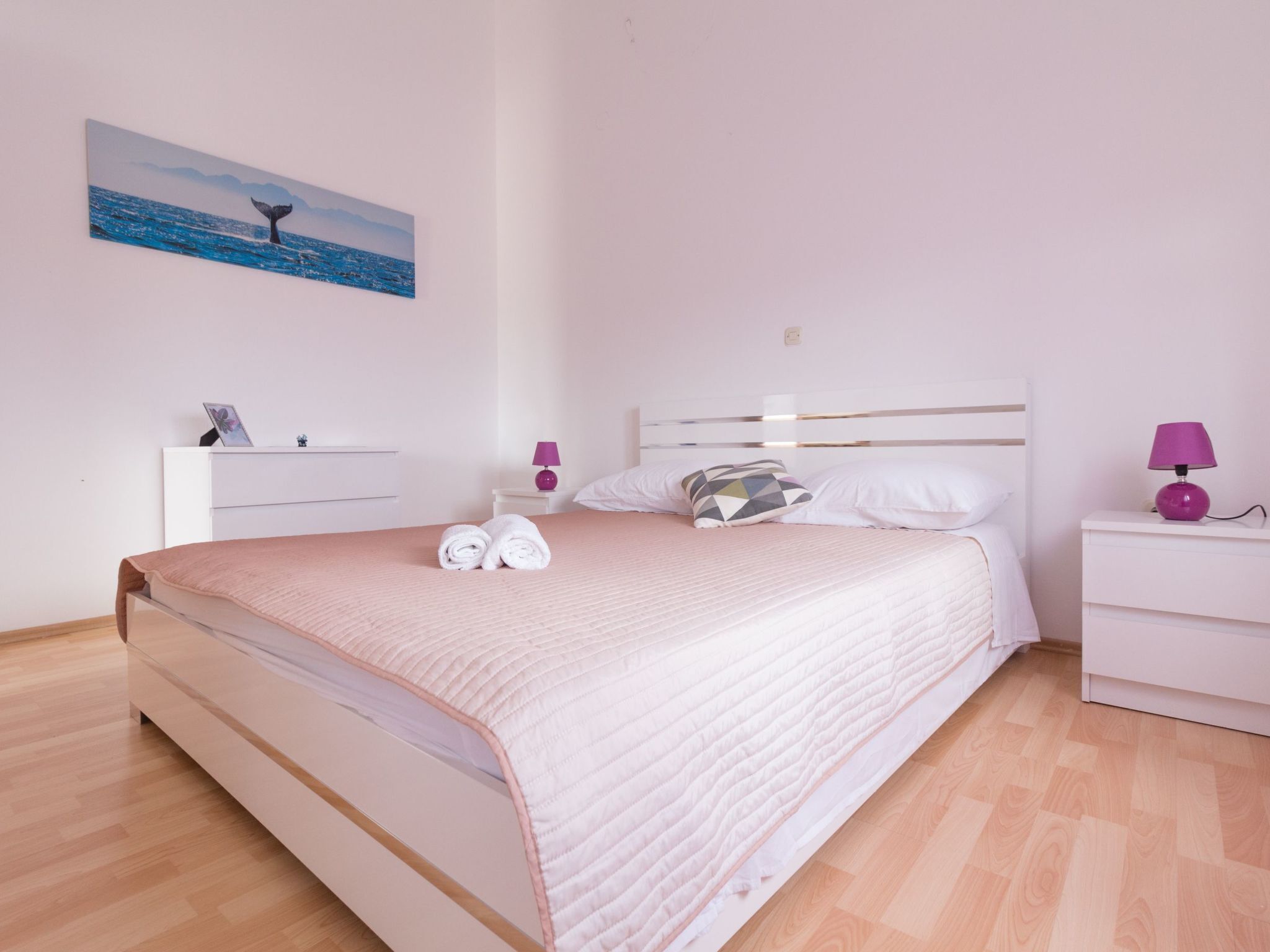 Ein ordentlich bezogenes Bett mit einer rosa Decke, einem weißen Kopfteil und einem Nachttisch mit einer violetten Lampe.