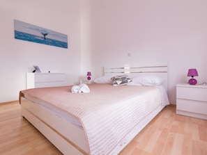 En pænt redt seng med et pink quilt, et hvidt sengebord og en lilla lampe.