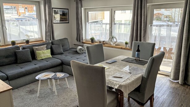 Ein modernes Wohnzimmer mit grauem Sofa, einem Esstisch mit Stühlen, großen Fenstern mit Vorhängen und Blick auf eine Straße.