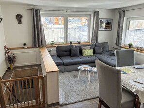 Una sala de estar moderna con un sofá seccional gris, una mesa de comedor con sillas y una escalera que sube a un piso superior.