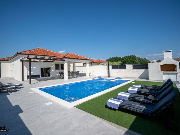 Ein moderner Außenpoolbereich mit einem klaren, blauen Pool, Liegestühlen und einer überdachten Terrasse.