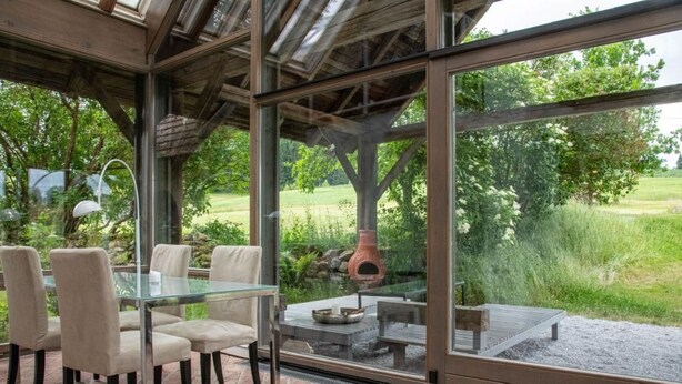 An outdoor dining area with a glass table and chairs, a brick floor, and a view of a garden and trees.