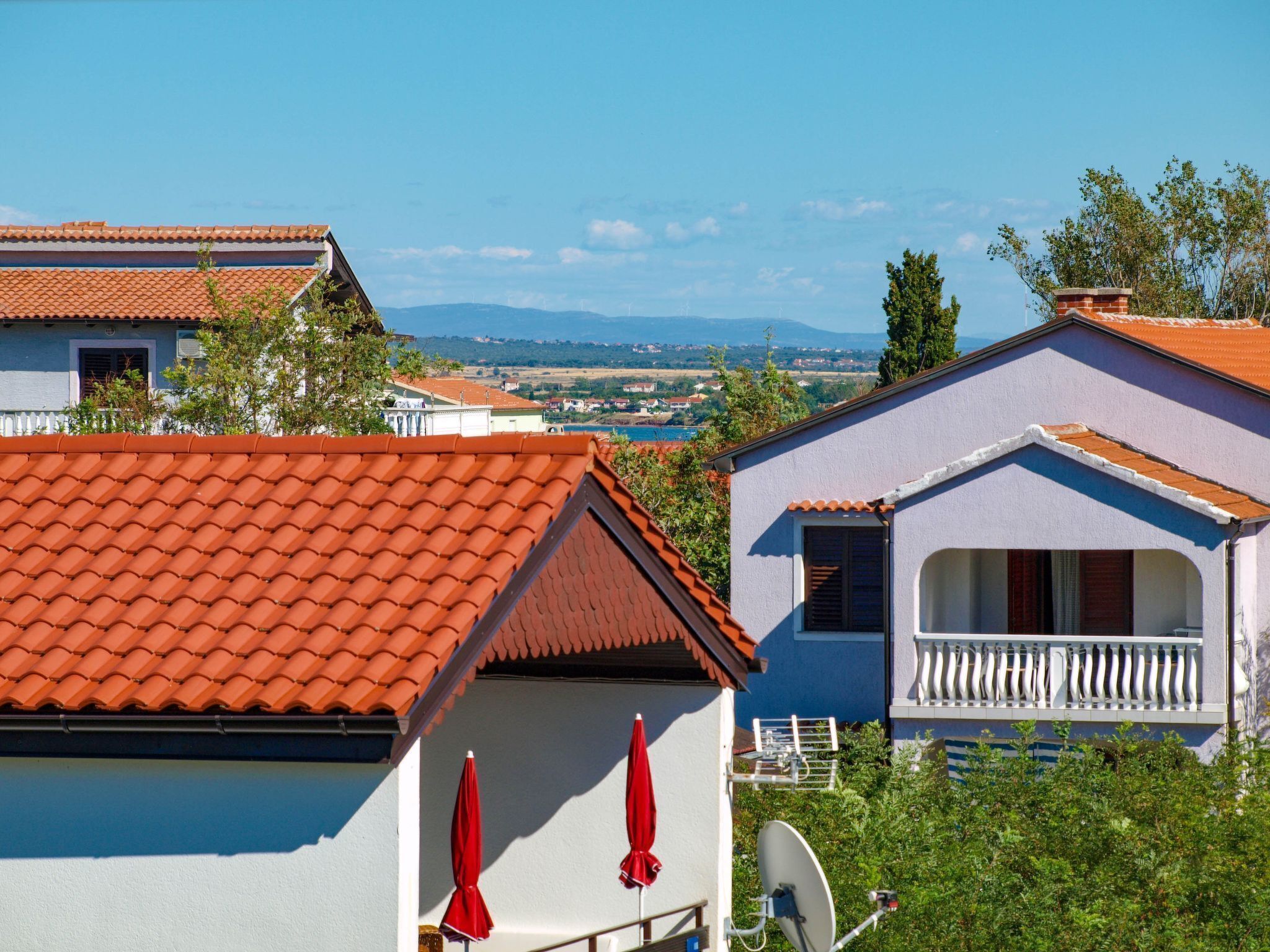 Eine Küstenvilla mit rotem Ziegeldach, einem Balkon mit weißen Geländern und Blick auf das Meer und ferne Berge.