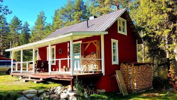 Une cabane rouge avec un porche, une pile de bois de chauffage et un mur en pierre.