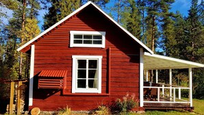 Une cabane en bois rouge, avec une fenêtre et une porte blanches, une véranda dotée de balustrades en bois et une vue dégagée sur la forêt environnante.