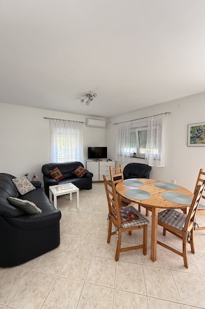 A living room with a black sofa, a wooden dining table with chairs, a TV, and a small white table. - Wohnung Vir in Strandnähe 01 - Agentur TA Leut by Interhome (Vir)