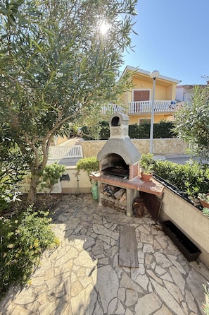 An outdoor stone patio with a wood-fired oven, surrounded by greenery and a yellow building in the background. - Wohnung Vir in Strandnähe 01 - Agentur TA Leut by Interhome (Vir)