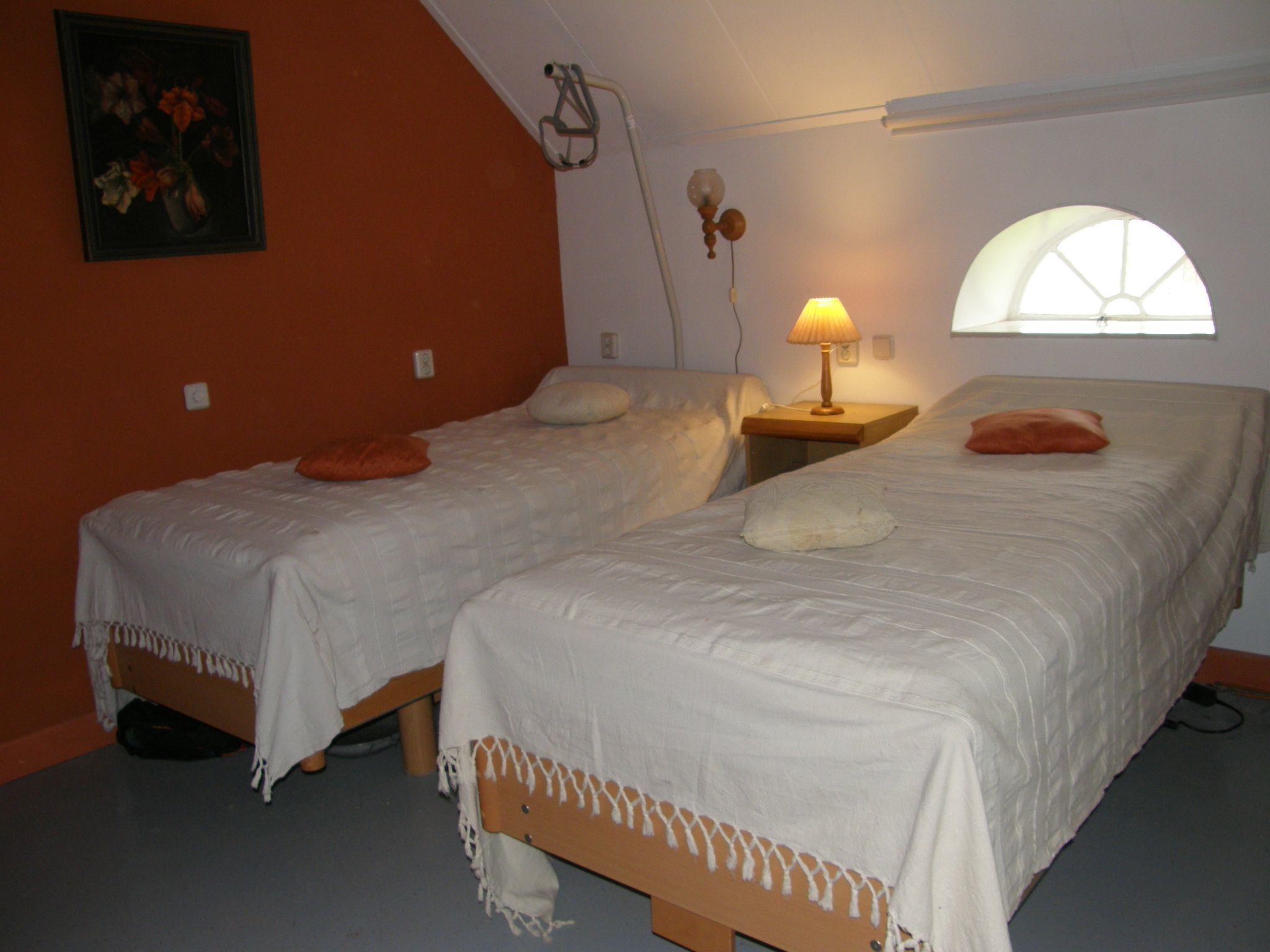 A bedroom with two single beds, a nightstand with a lamp, a wall-mounted heater, and a window with a view.