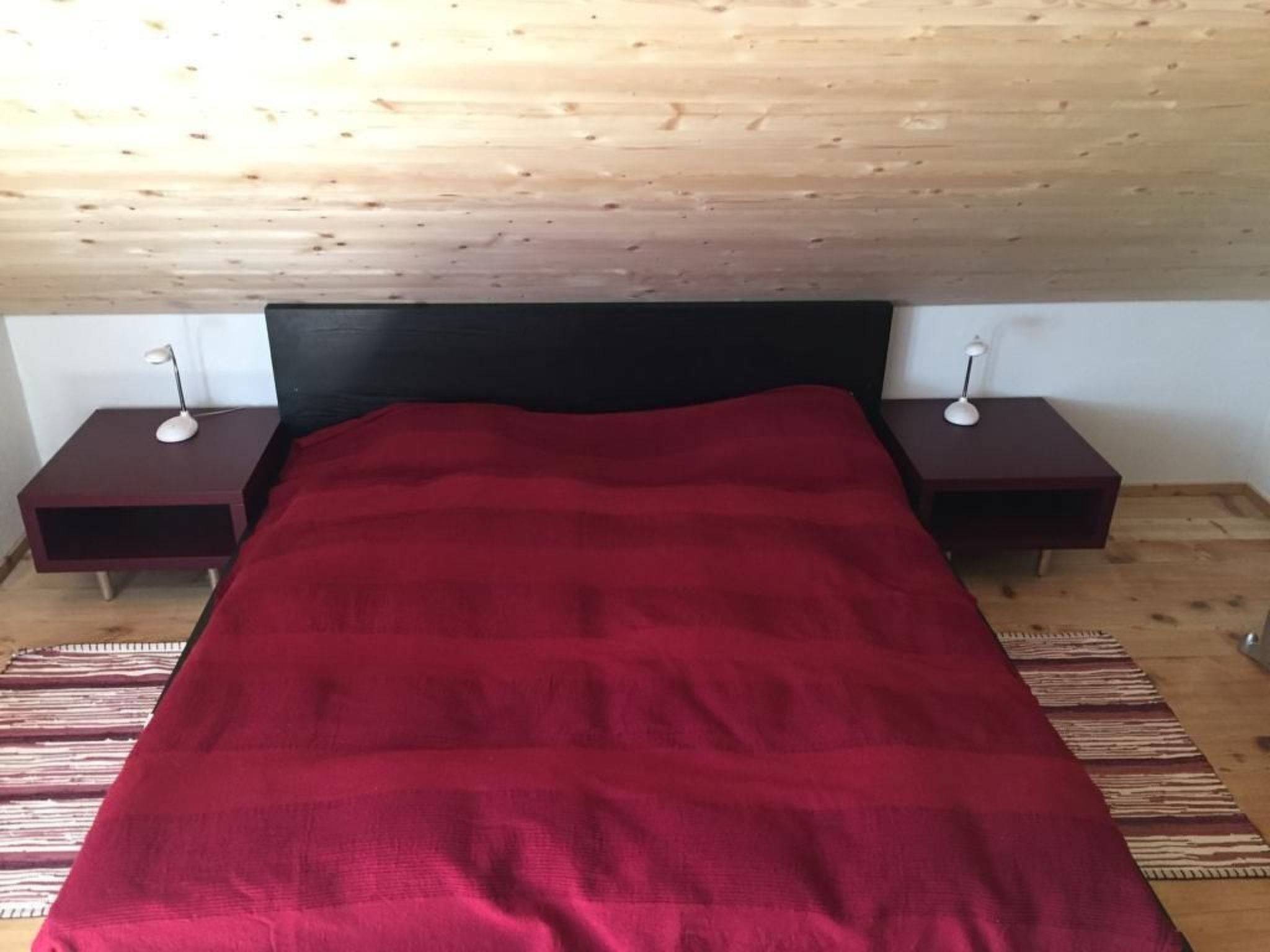 Ein Schlafzimmer mit rotem Bettbezug, einem Holz-Kopfteil, zwei Nachttischen mit Lampen und einem gemusterten Teppich.