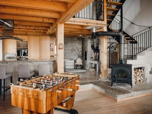 Un intérieur chaleureux et rustique, avec un salon doté de planchers en bois, une table de baby-foot, une cheminée en pierre et un escalier menant à un étage supérieur.