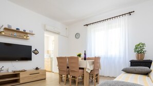 Una sala moderna con televisor de pantalla plana, muebles de madera, una mesa de comedor con sillas, una cama con ropa de cama a rayas y una planta en maceta.