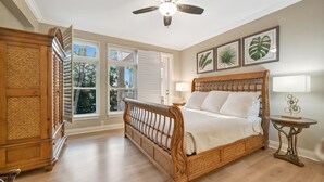 Een slaapkamer met een houten bed, een plafondventilator, een raam met luiken en plantenkunst in een lijst aan de wand.