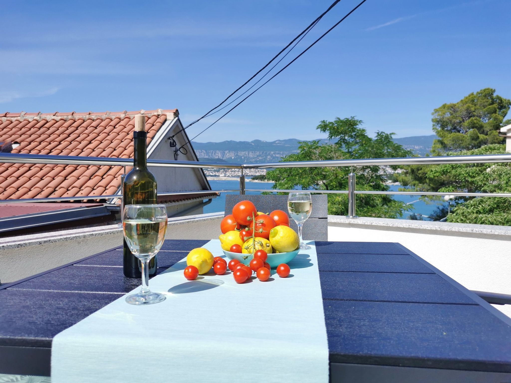 Een tafel met een fles witte wijn, een glas en een schaal tomaten en citroenen op een balkon met uitzicht op een kustgebied, een betegeld dak en een zeezicht.