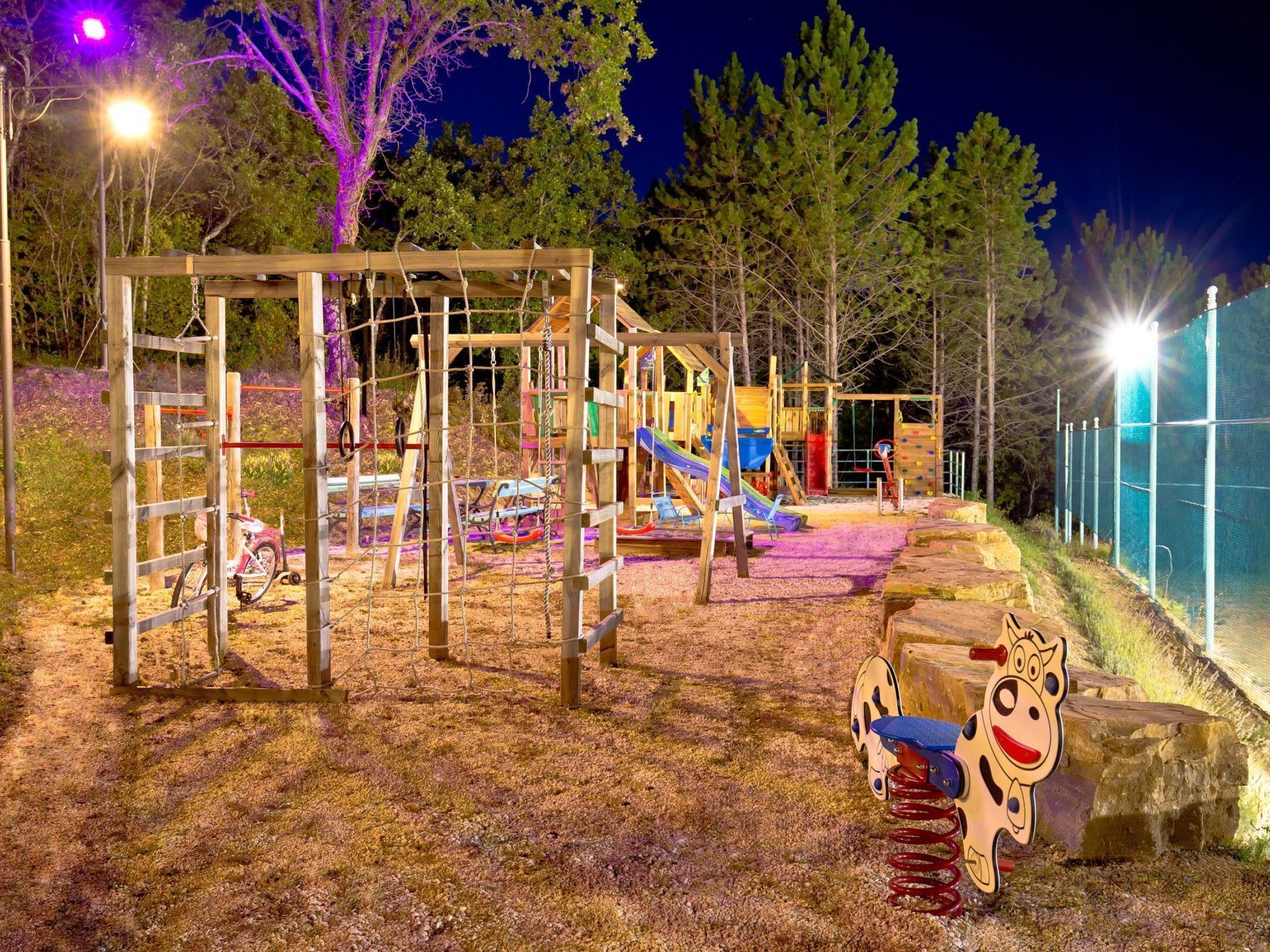Ein Spielplatz mit verschiedenen Spielgeräten, darunter eine Rutsche und Klettergerüste, umgeben von Bäumen und einem eingezäunten Bereich.