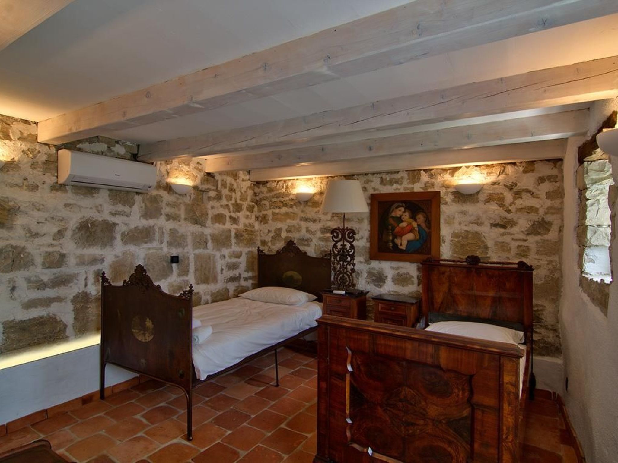 A rustic bedroom with exposed stone walls, a wooden ceiling, and a tiled floor. There are two wooden beds with headboards, a bedside table, and a wall-mounted lamp.