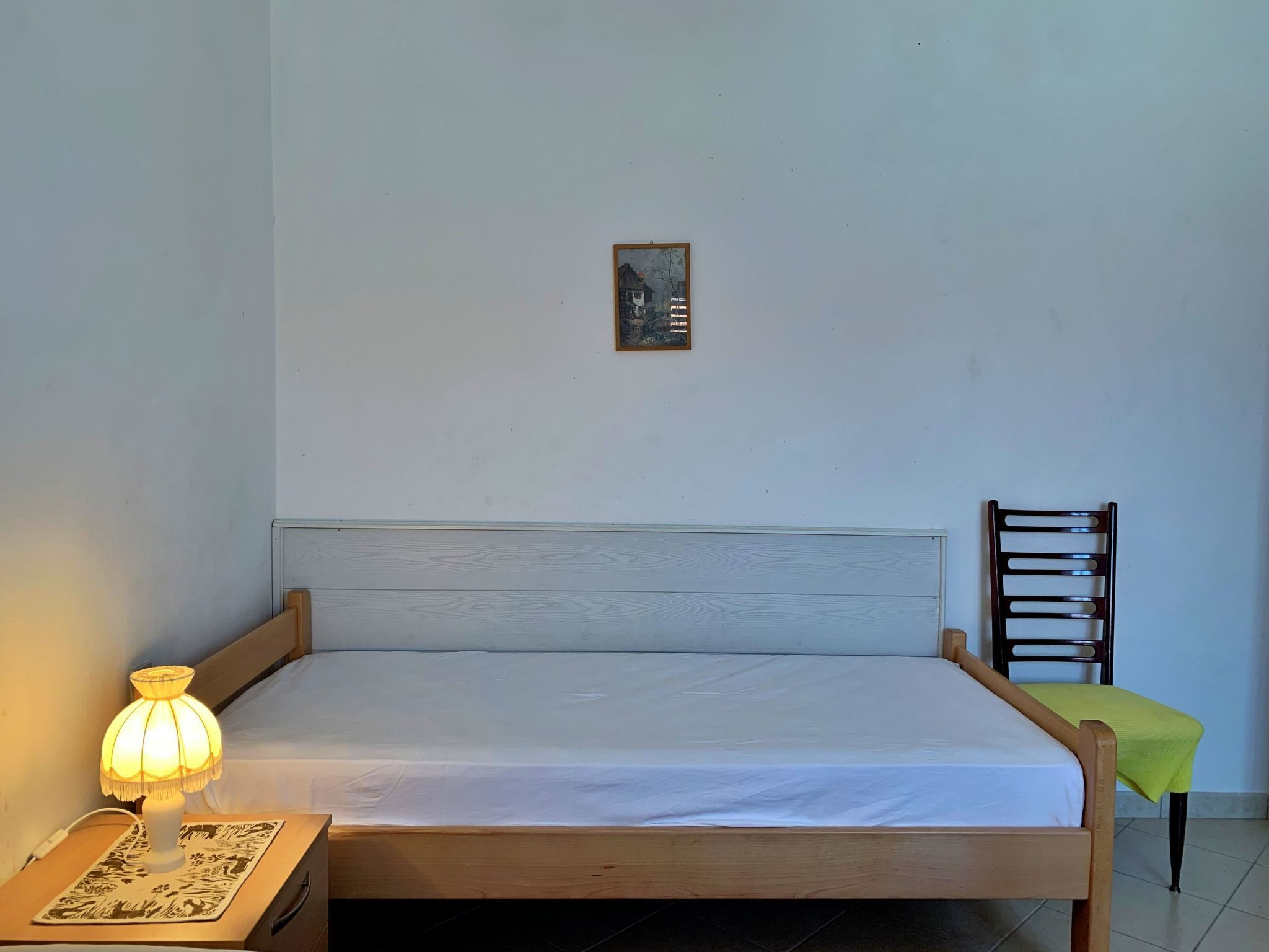 Un lit simple avec un cadre en bois et du linge de lit blanc, une lampe de chevet, un petit tableau encadré accroché au mur et une chaise recouverte d’un tissu vert.