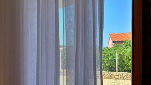 A window with sheer white curtains, a stone patio, and a red-roofed building in the background. - Wohnung Volaric by Interhome (Polje)