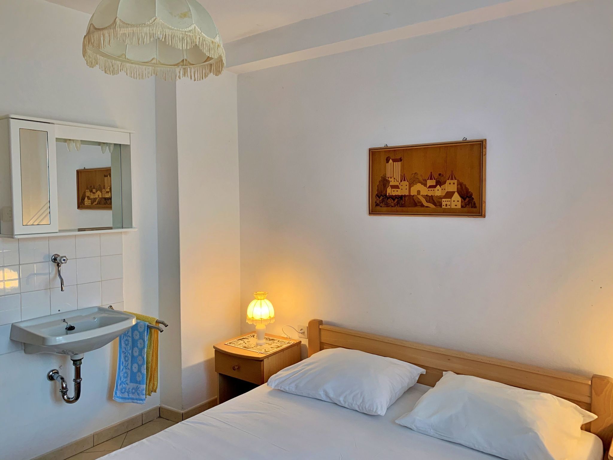 A hotel room with a bed, a bedside table with a lamp, a wall-mounted sink, a towel, and a painting of a town on the wall.