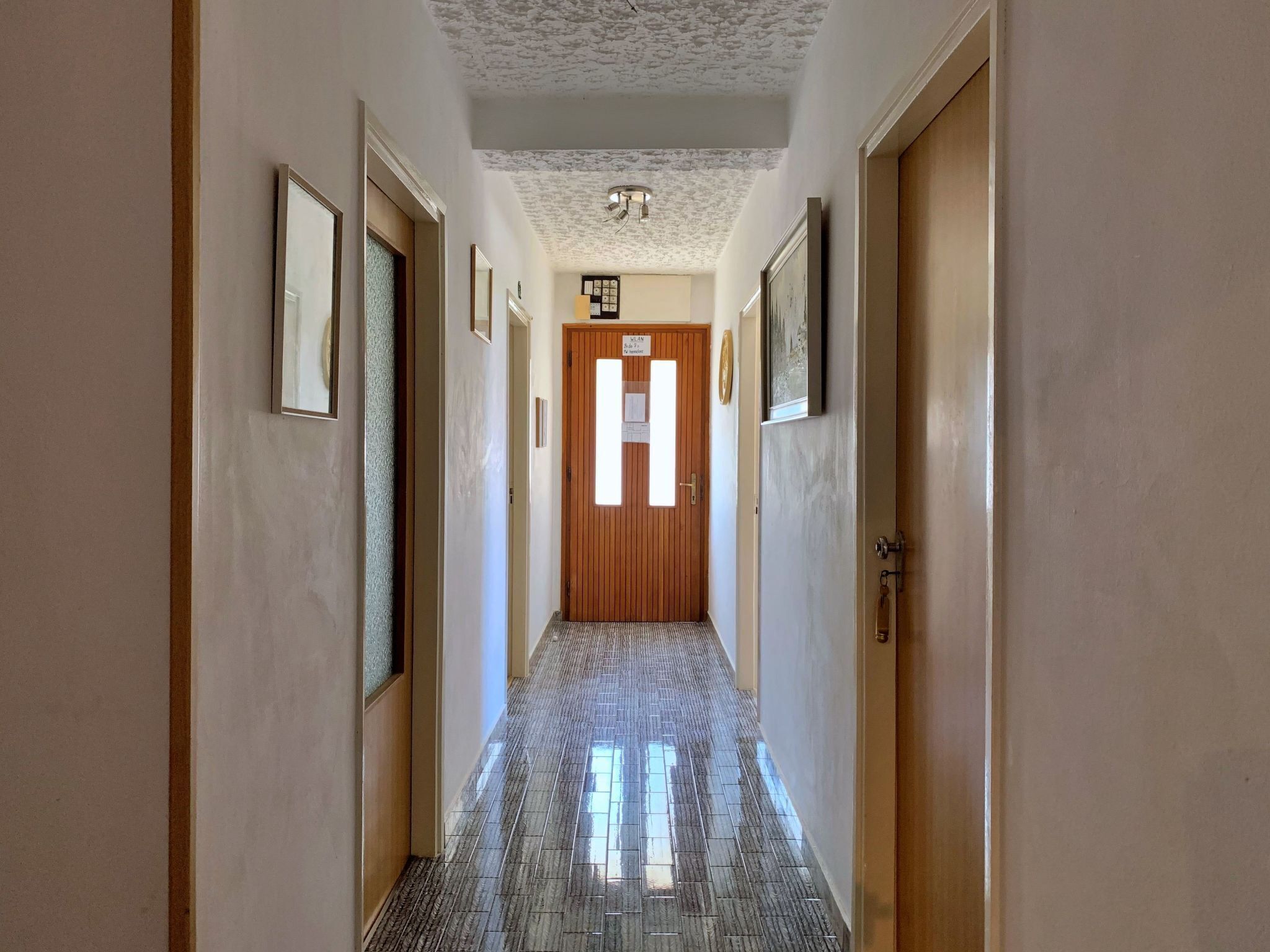 A hallway with a wooden door at the end, framed pictures on the walls, and a tiled floor.