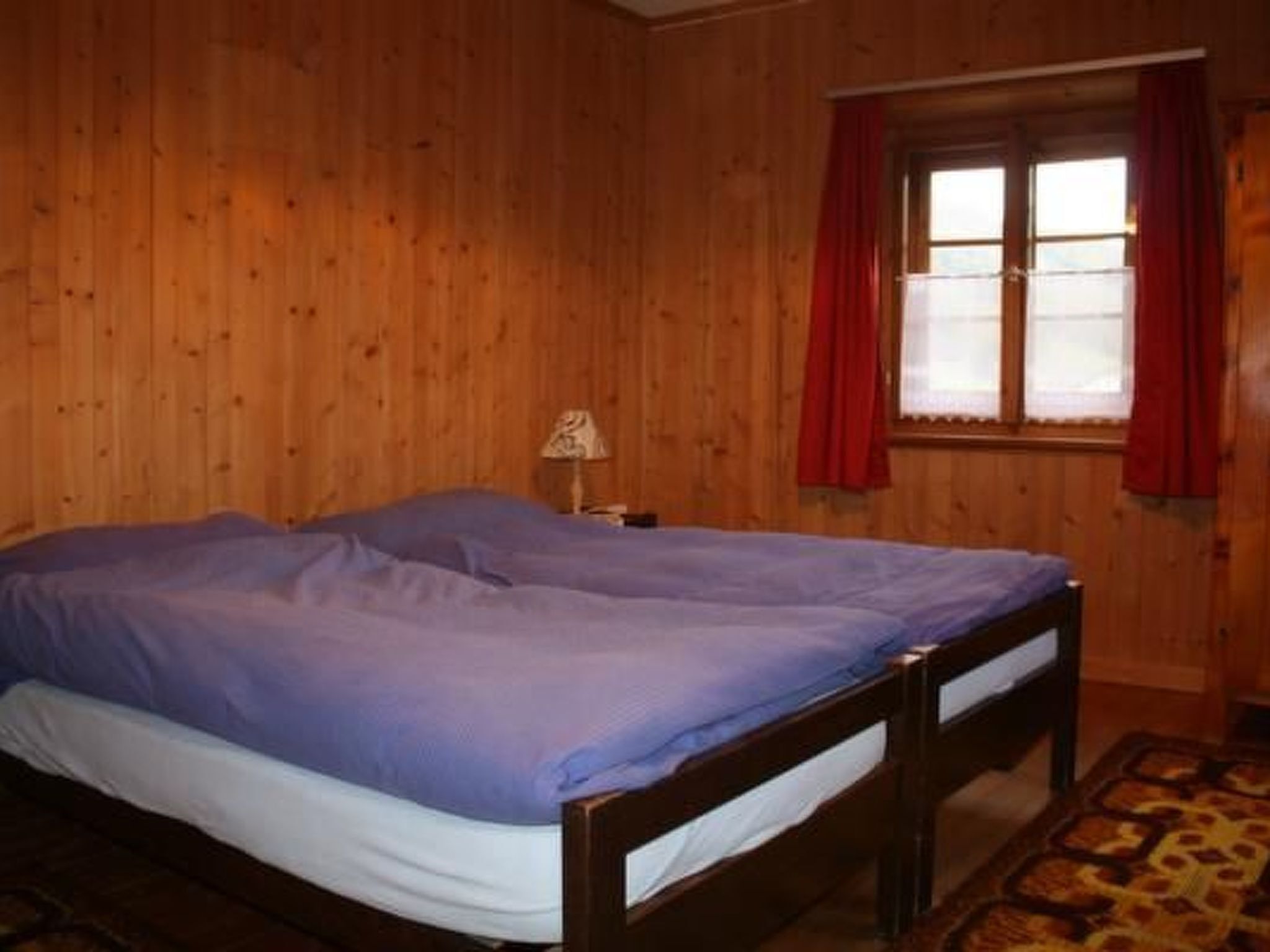 Una habitación con paredes de madera, una cama con colcha azul, una mesita de noche con lámpara y una ventana con cortinas rojas.